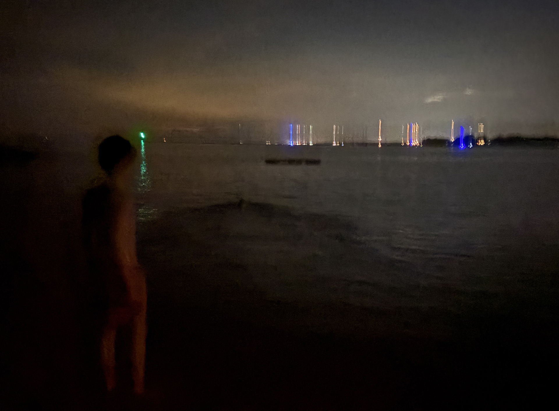 small boat lights in the distance as a figure wades into the water, like a ghost, towards peaceful dark waves and the veil of night